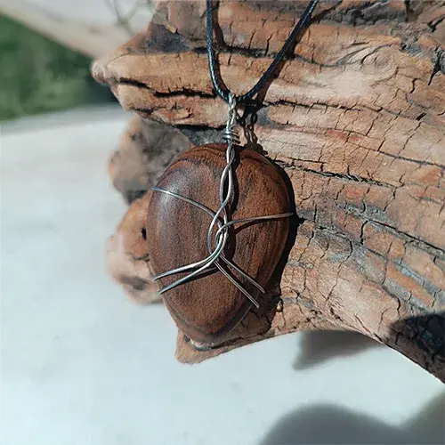 Pendentif Étreinte d’Acier en noyer, enlacé de fils d’acier inoxydable 316L, présenté sur un support en bois flotté.