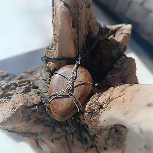 Pendentif Étreinte d’Acier en noyer et fil d’acier inoxydable 316L, photographié de trois quarts sur un support en bois flotté.