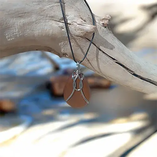 Pendentif Noyer Enlacé en noyer et fil d’acier inoxydable 316L, suspendu par son cordon noir à une branche de bois flotté.